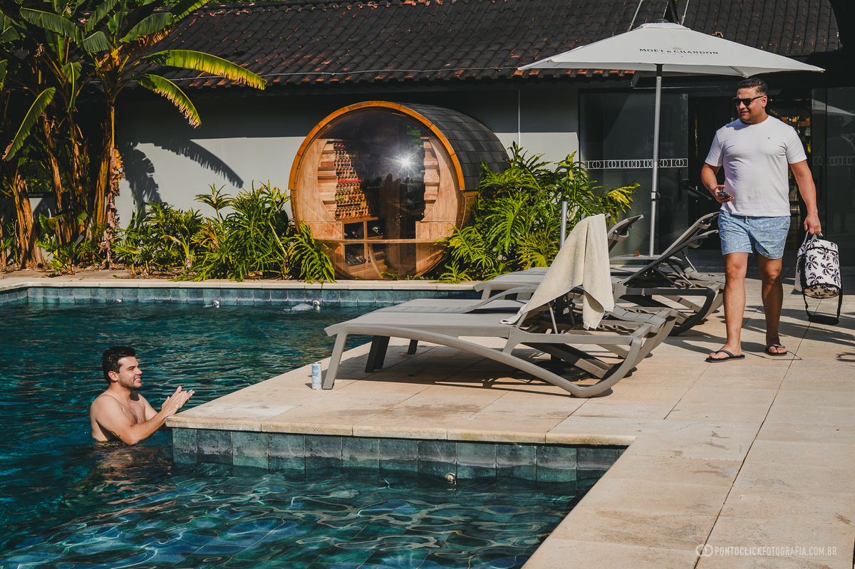 Um homem à beira da piscina conversa com outro que está na água, em uma cena espontânea do dia do casamento. Fotógrafo de casamento Pontoclick Fotografia.