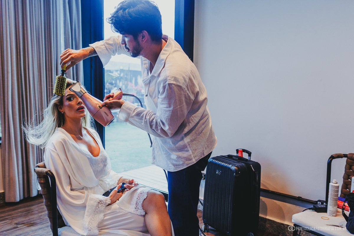 O cabeleireiro trabalha no penteado da noiva enquanto ela aguarda sentada em frente à janela. Fotógrafo de casamento Pontoclick Fotografia.