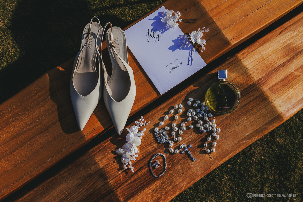 Sapatos brancos, joias, perfume e convite estão dispostos sobre a mesa em uma fotografia dos detalhes da noiva. Fotógrafo de casamento Pontoclick Fotografia.