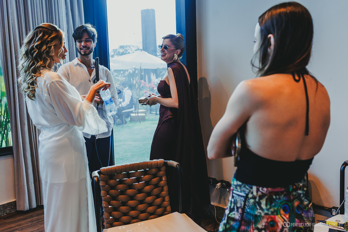 A noiva, vestida com um robe branco, conversa com outras pessoas em uma sala iluminada durante os preparativos. Fotógrafo de casamento Pontoclick Fotografia.