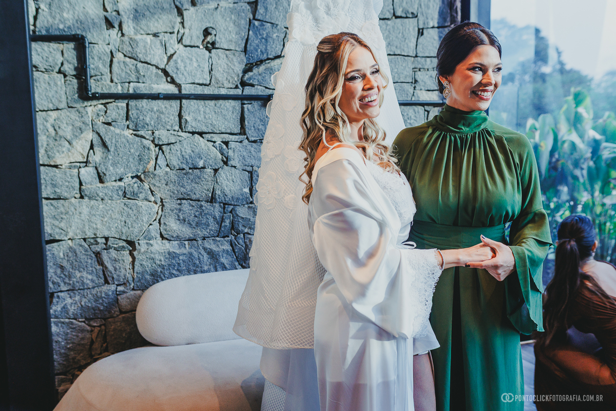 A noiva de robe branco sorri ao lado de uma madrinha de vestido verde, com o vestido de casamento ao fundo. Fotógrafo de casamento Pontoclick Fotografia