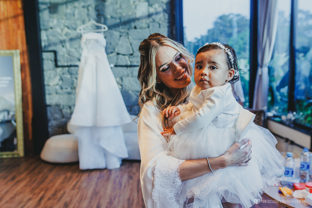 A noiva segura uma criança de vestido branco durante o making of, com o vestido de casamento ao fundo. Fotógrafo de casamento Pontoclick Fotografia