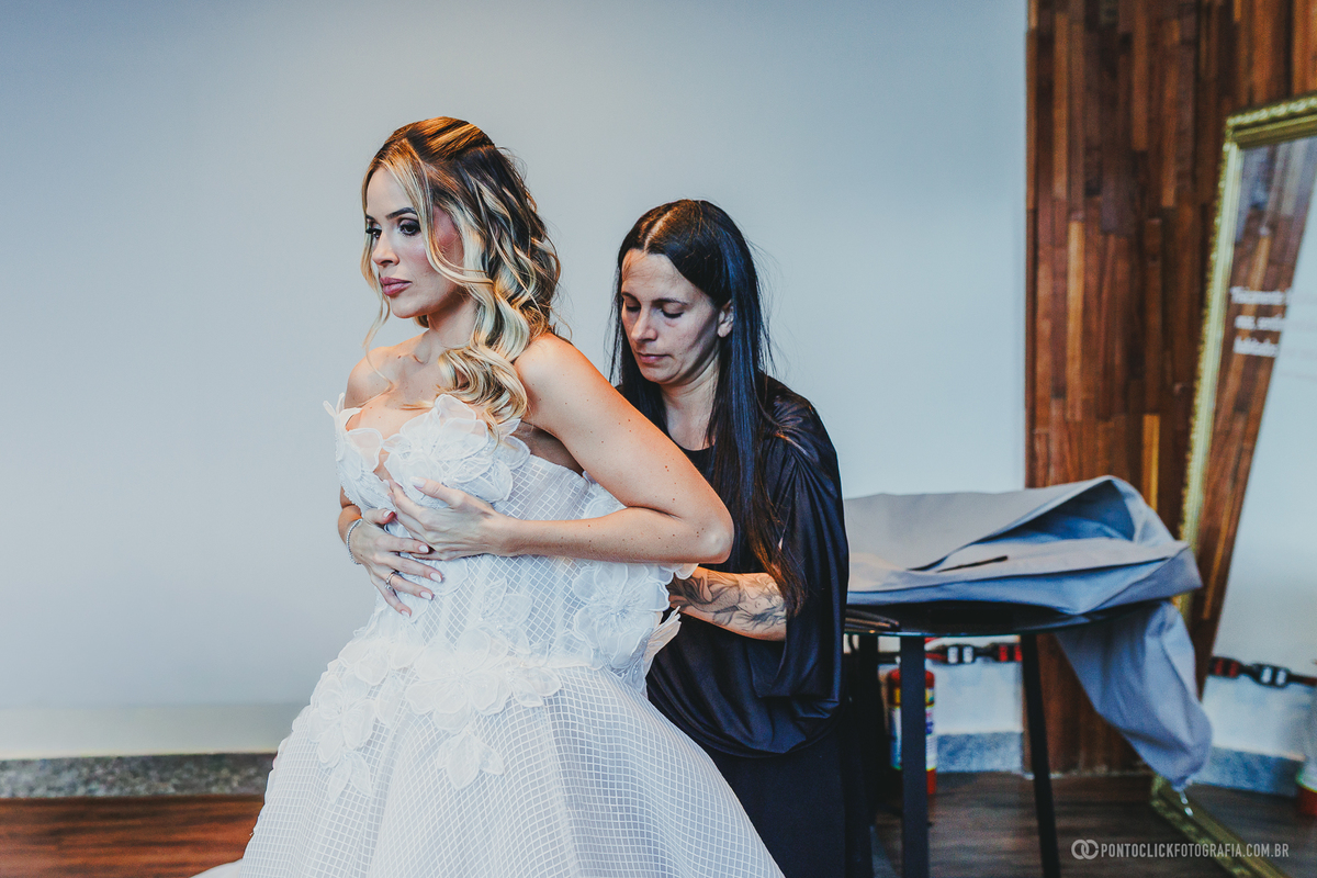 A noiva veste o vestido branco com aplicações florais enquanto recebe ajuda para ajustá-lo. Fotógrafo de casamento Pontoclick Fotografia