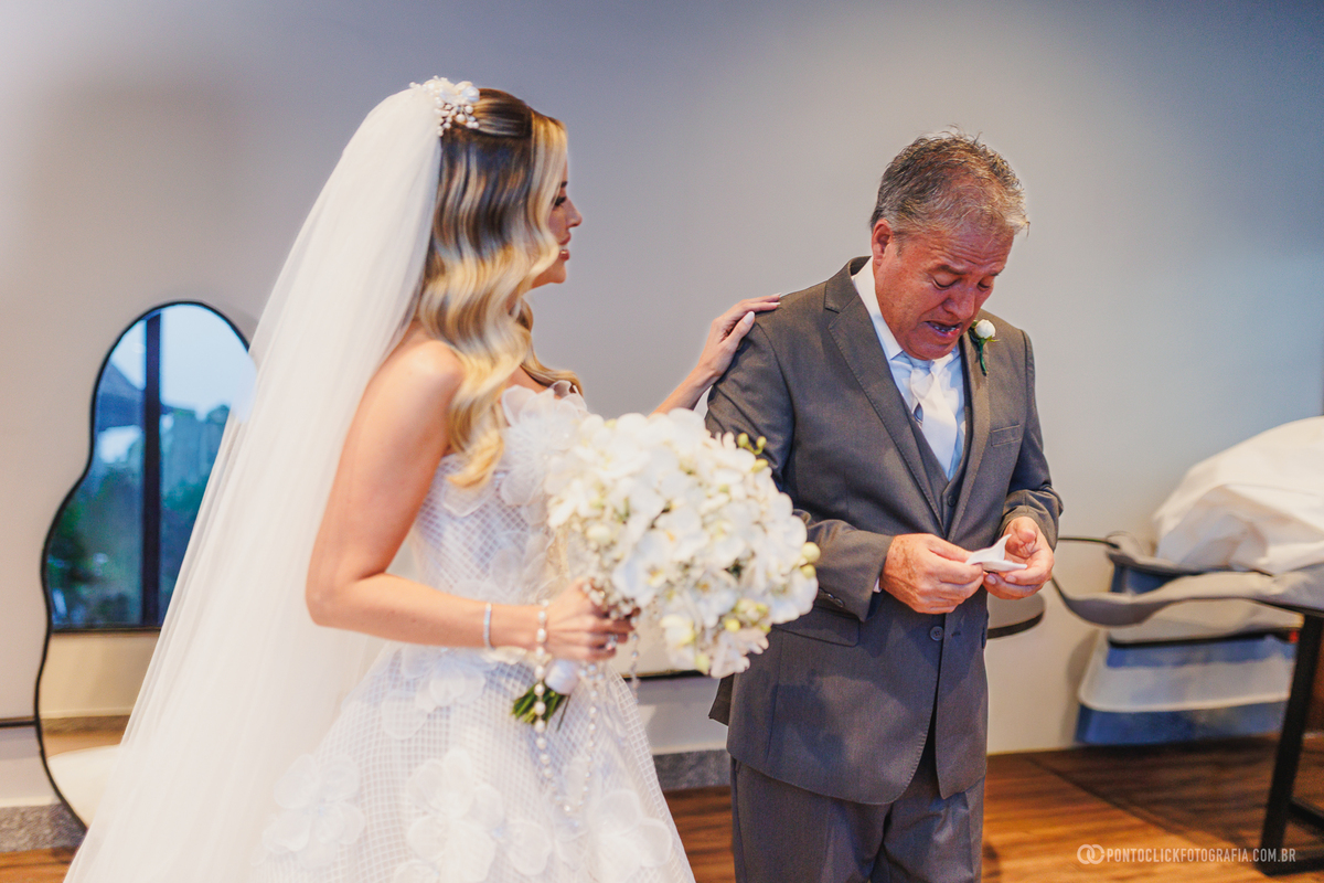 A noiva, com vestido, véu e buquê, acompanha o pai de terno que lê uma mensagem durante os preparativos do casamento. Fotógrafo de casamento Pontoclick Fotografia.