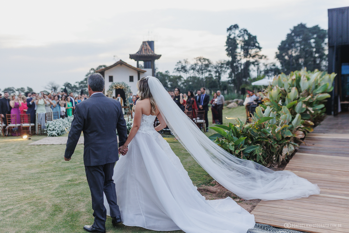 A noiva caminha de mãos dadas com o pai de terno em direção à cerimônia ao ar livre, com o longo véu estendido atrás dela. Fotógrafo de casamento Pontoclick Fotografia.