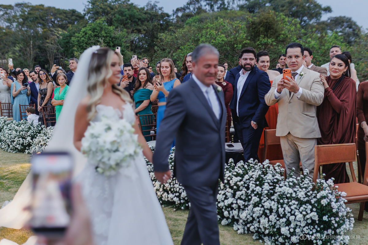 A noiva caminha pela cerimônia ao ar livre acompanhada por um homem de terno, enquanto os convidados registram sua entrada. Fotógrafo de casamento Pontoclick Fotografia.