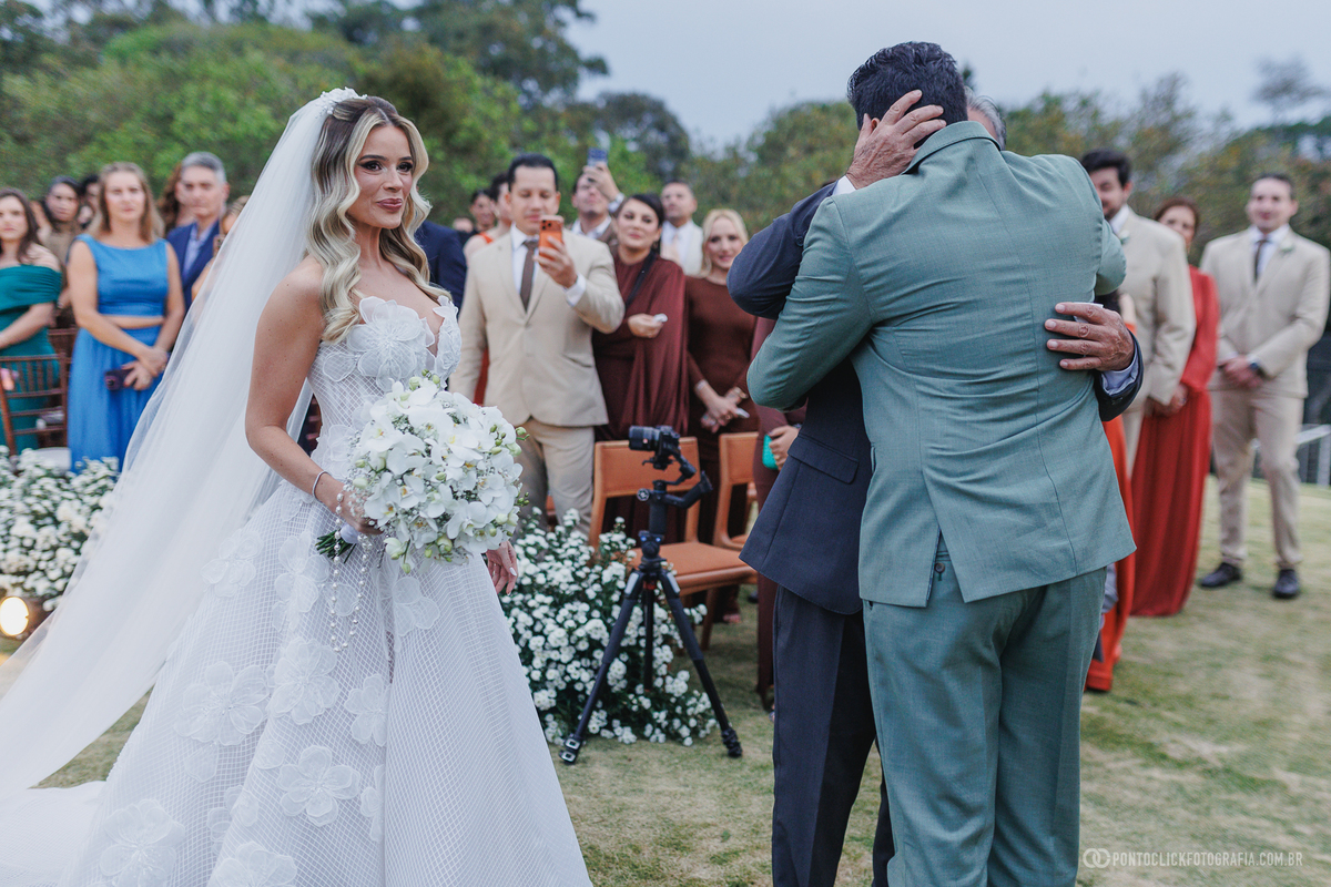 A noiva segura o buquê enquanto o noivo abraça um homem de terno durante a cerimônia. Fotógrafo de casamento Pontoclick Fotografia.