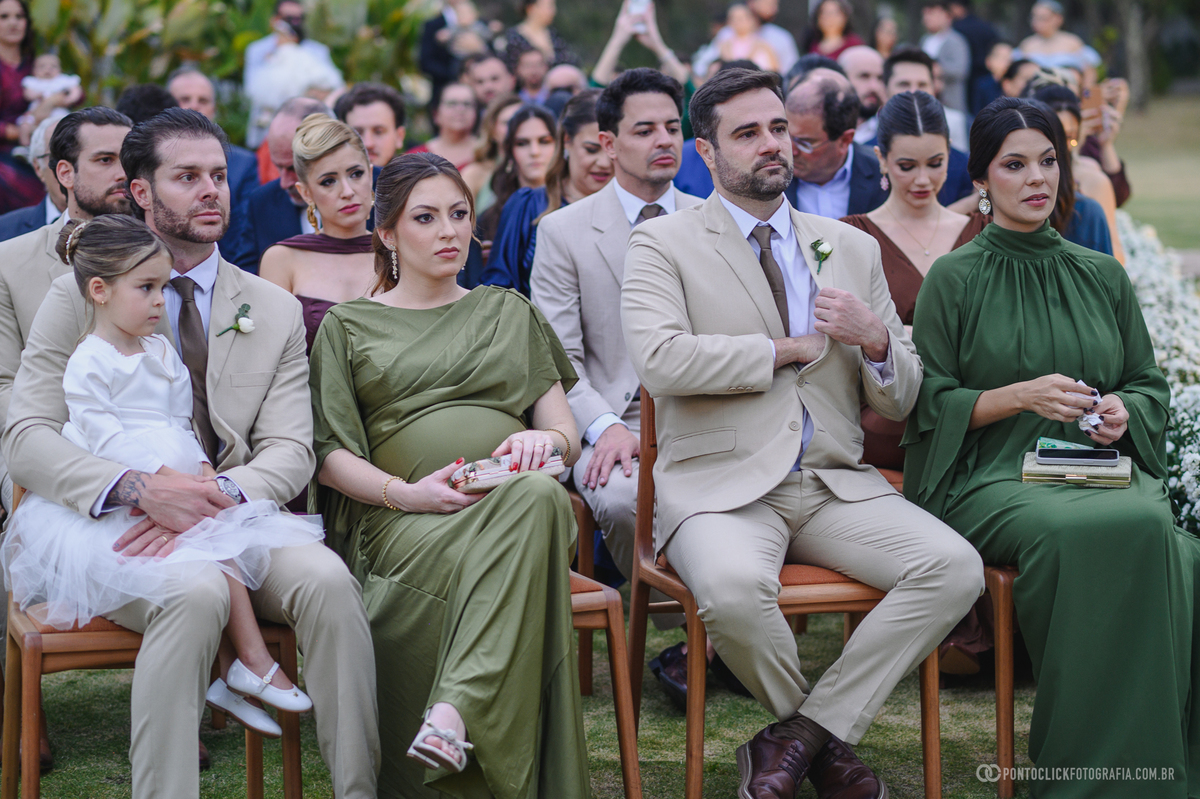 Convidados em trajes claros e verdes acompanham a cerimônia, com uma criança de vestido branco na primeira fileira. Fotógrafo de casamento Pontoclick Fotografia.
