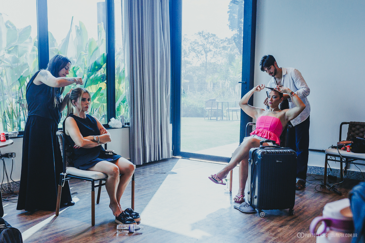Duas mulheres têm o cabelo preparado diante das janelas, em meio à movimentação do making of. Fotógrafo de casamento Pontoclick Fotografia