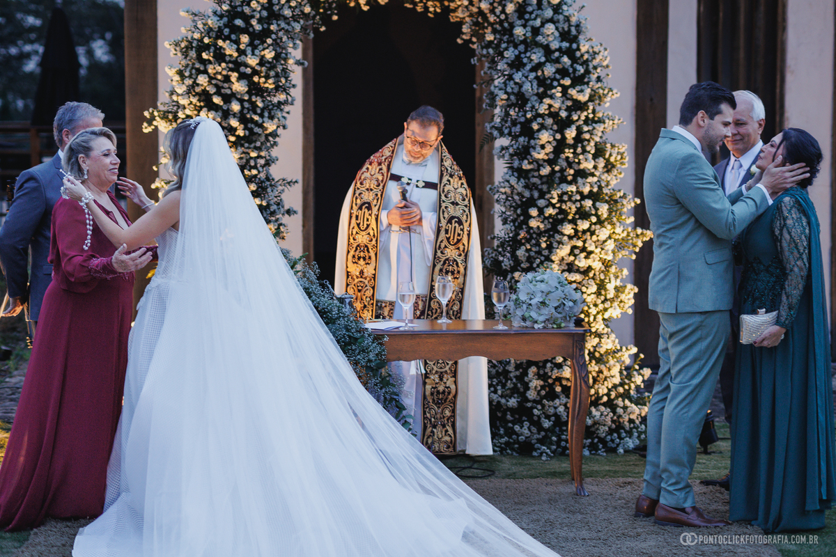 Os noivos cumprimentam os pais  diante do celebrante e de um arco de flores durante a cerimônia de casamento. Fotógrafo de casamento Pontoclick Fotografia