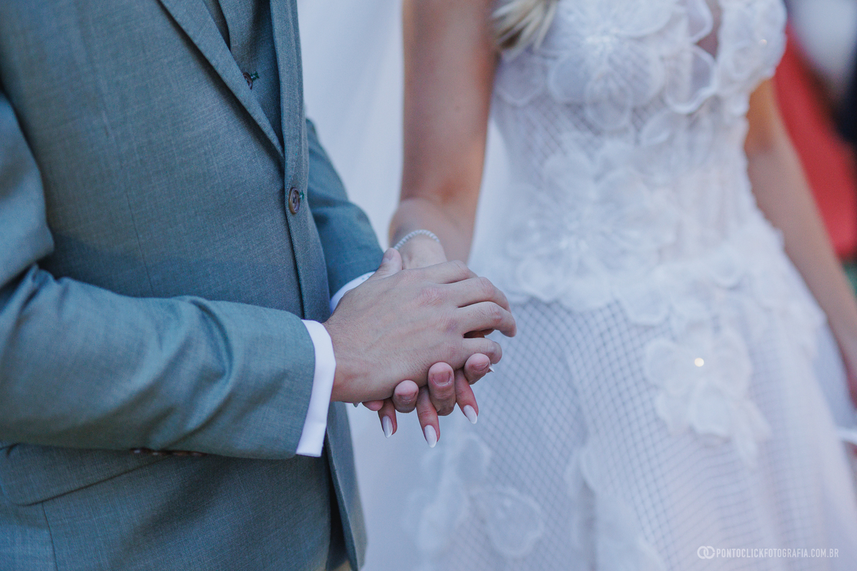 Detalhe das mãos dos noivos unidas durante a cerimônia, com o terno verde e o vestido branco em destaque. Fotógrafo de casamento Pontoclick Fotografia