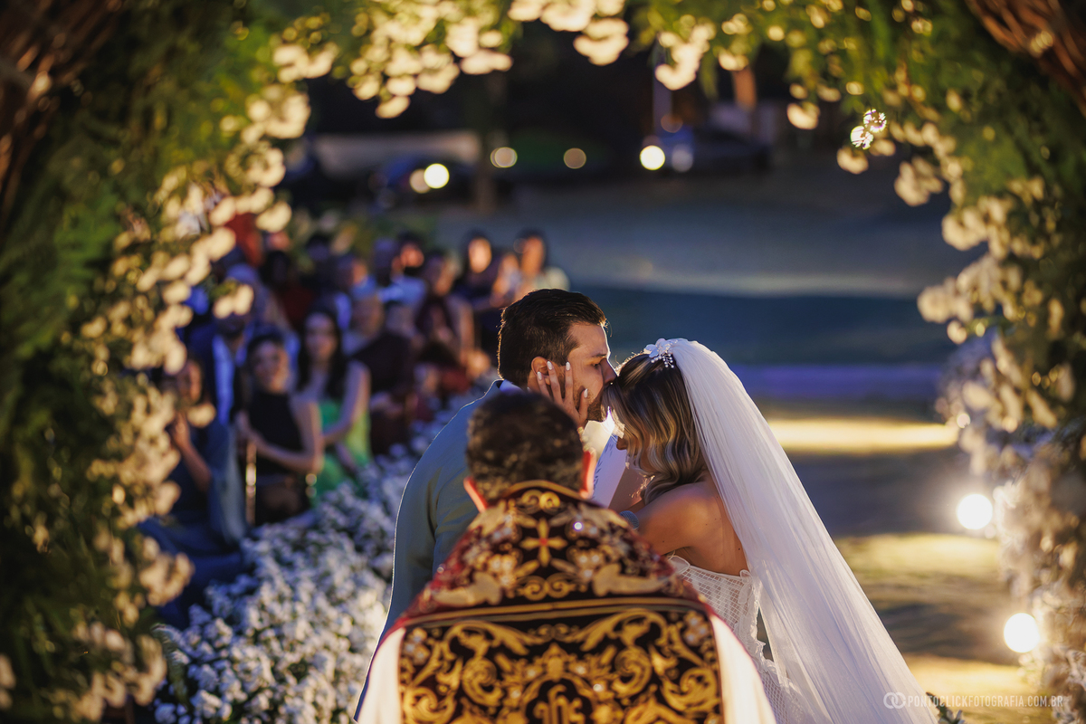 Os noivos se beijam sob um arco de flores iluminado durante a cerimônia ao entardecer. Fotógrafo de casamento Pontoclick Fotografia.
