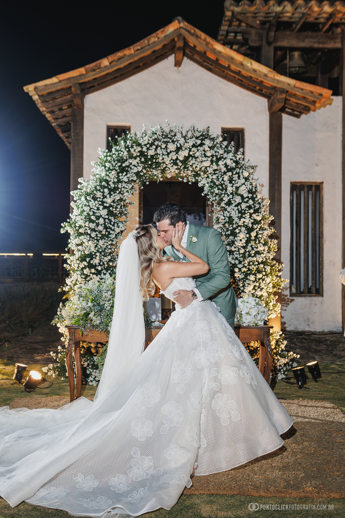 Os noivos se beijam diante do arco de flores iluminado, com o vestido e o longo véu da noiva em destaque. Fotógrafo de casamento Pontoclick Fotografia