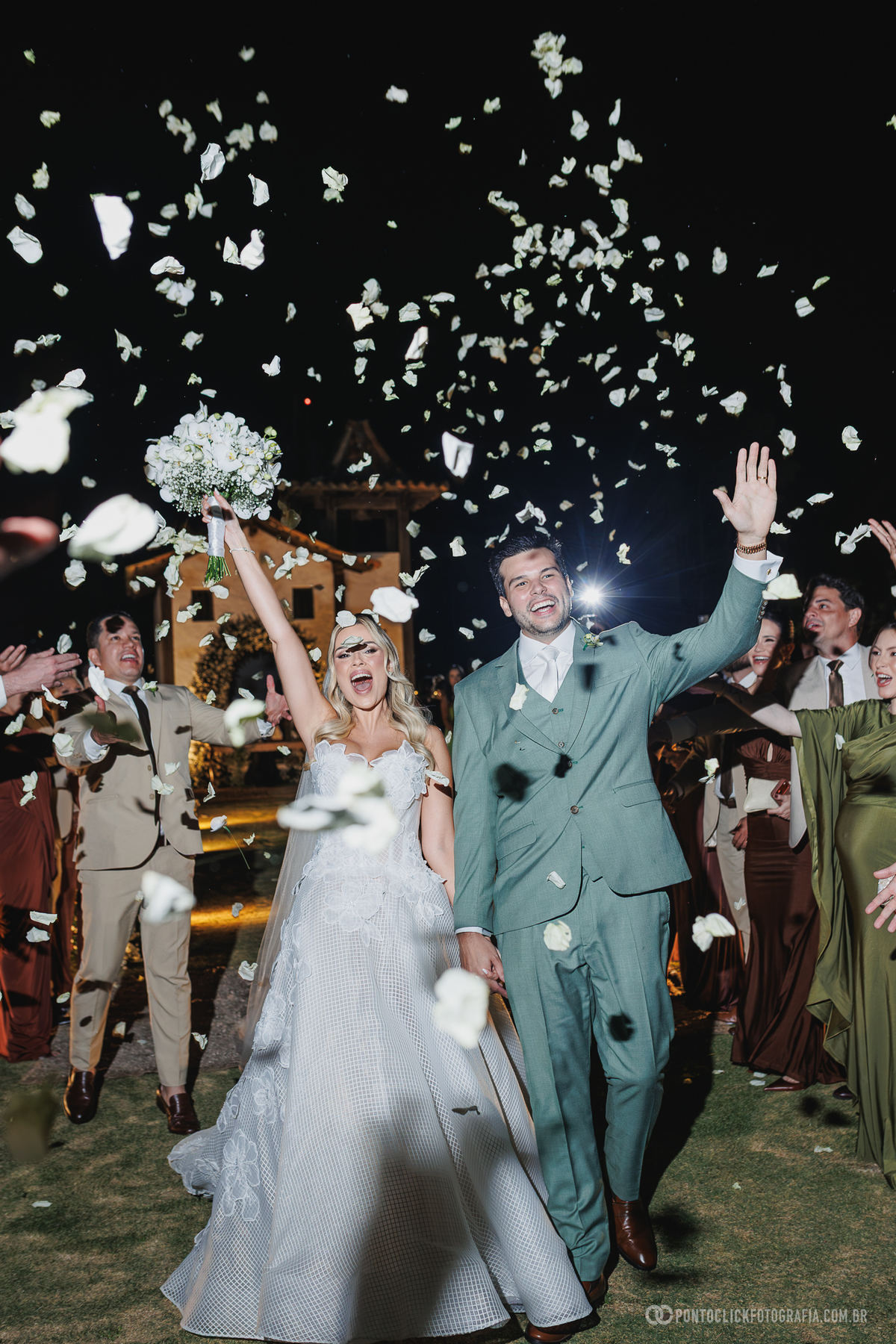Os noivos saem da cerimônia de mãos dadas, comemorando sob uma chuva de pétalas brancas lançadas pelos convidados. Fotógrafo de casamento Pontoclick Fotografia.