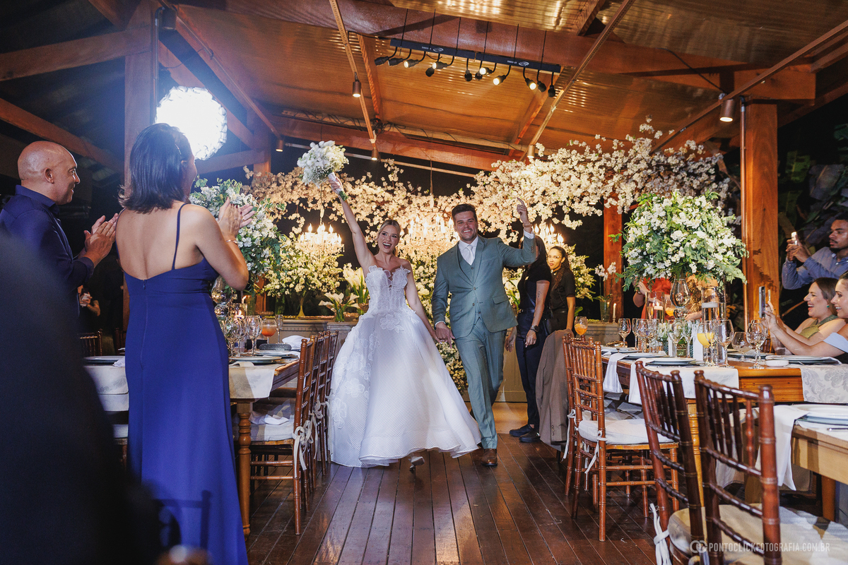 Os noivos entram juntos na recepção de casamento, entre mesas decoradas com flores e convidados que os aplaudem. Fotógrafo de casamento Pontoclick Fotografia