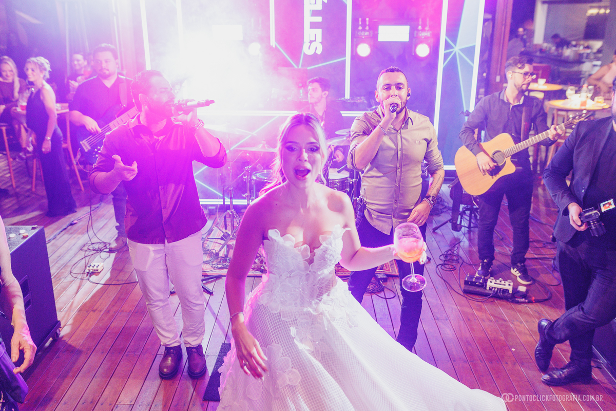 A noiva dança com uma taça na mão diante da banda que anima a festa de casamento. Fotógrafo de casamento Pontoclick Fotografia.