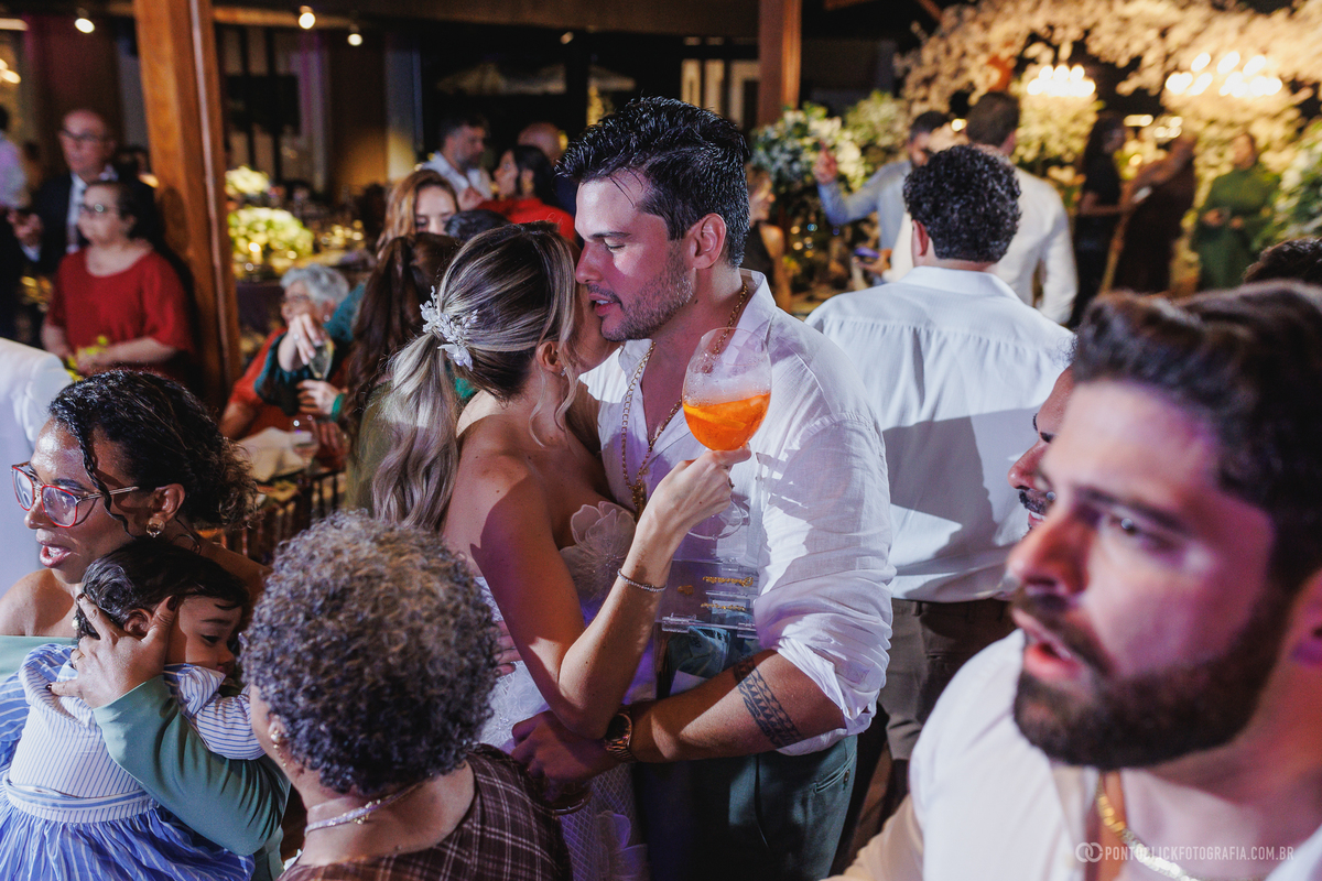 Os noivos se abraçam durante a festa de casamento, entre convidados e taças erguidas. Fotógrafo de casamento Pontoclick Fotografia.