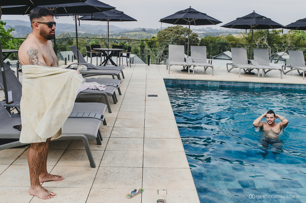 Um homem segura uma toalha à beira da piscina enquanto noivo permanece na água, em um momento descontraído. Fotógrafo de casamento Pontoclick Fotografia