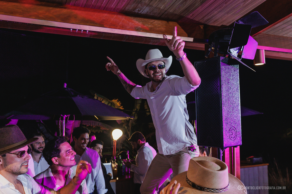 Um convidado de chapéu comemora de braços erguidos em meio à pista de dança. Fotógrafo de casamento Pontoclick Fotografia