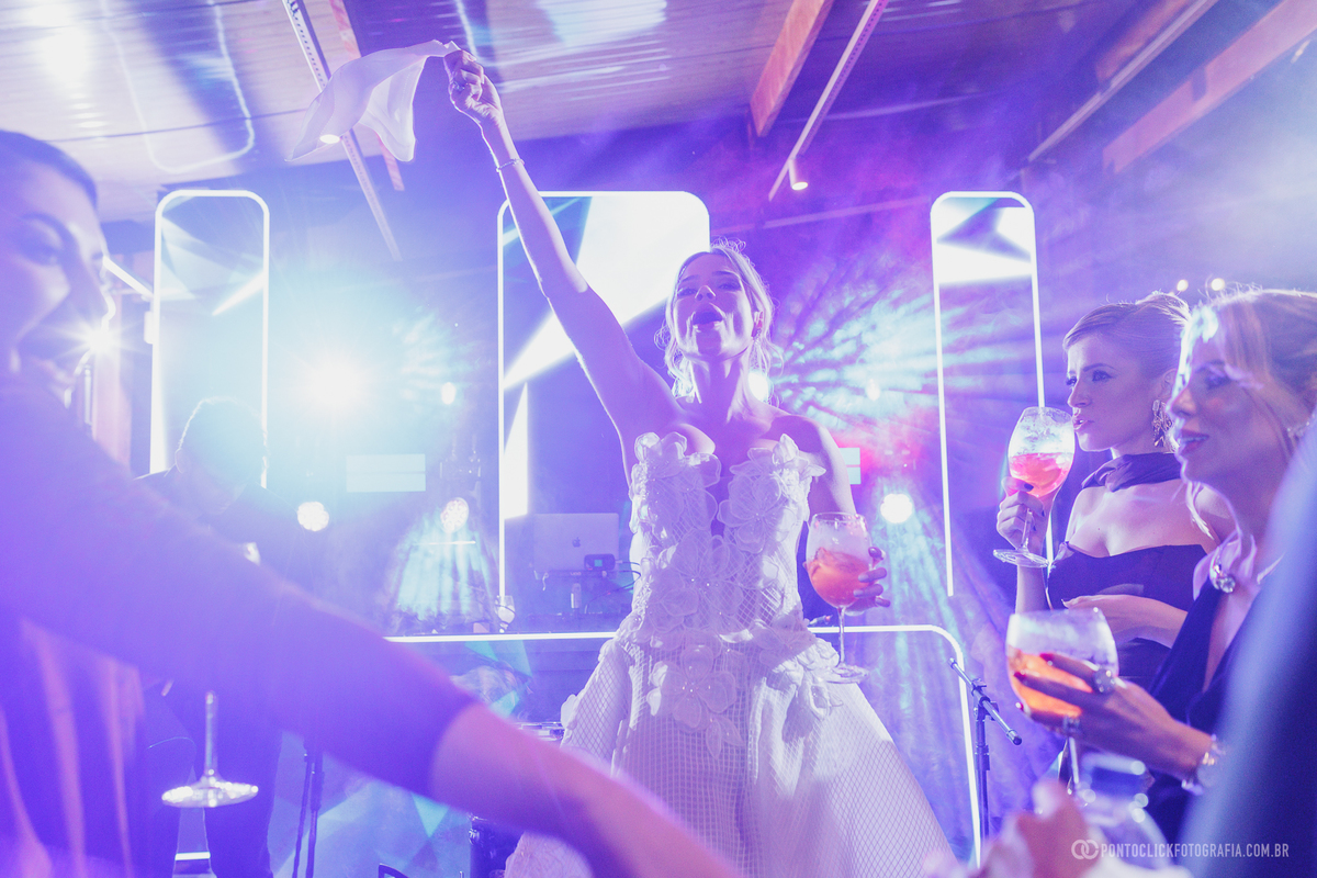 A noiva dança com convidadas, erguendo um tecido branco e segurando uma taça sob as luzes da festa. Fotógrafo de casamento Pontoclick Fotografia.