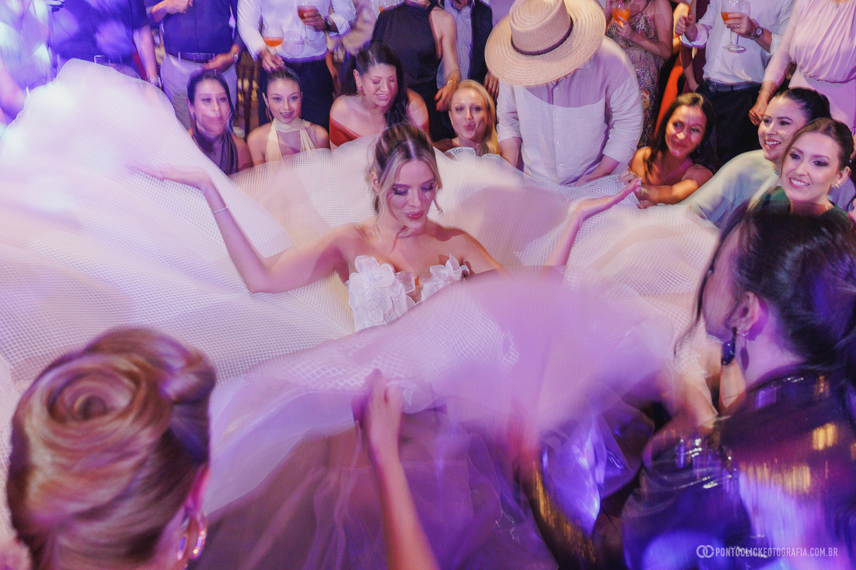 Noiva dança enquanto convidadas movimentam a saia do vestido na pista, sob luzes coloridas. Fotógrafo de casamento Pontoclick Fotografia.