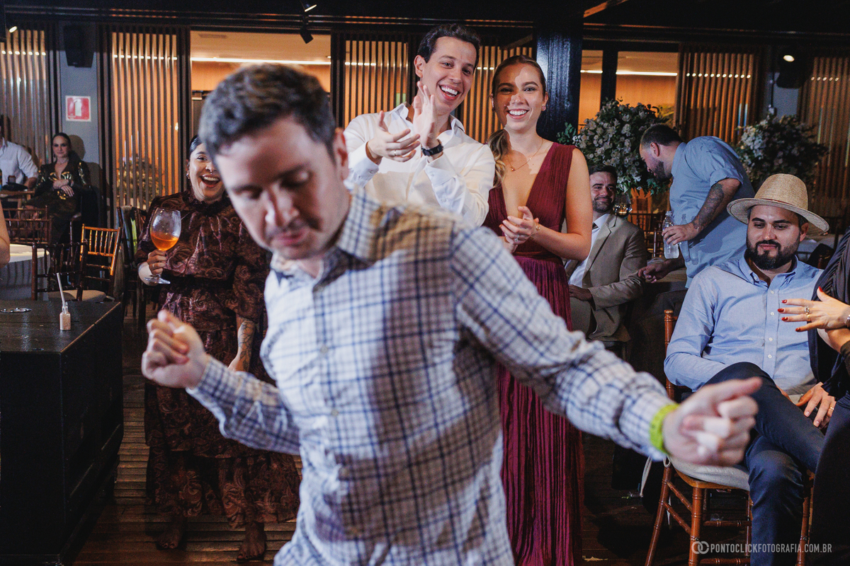 Convidado dança em primeiro plano enquanto amigos sorriem e aplaudem na festa de casamento. Fotógrafo de casamento Pontoclick Fotografia.