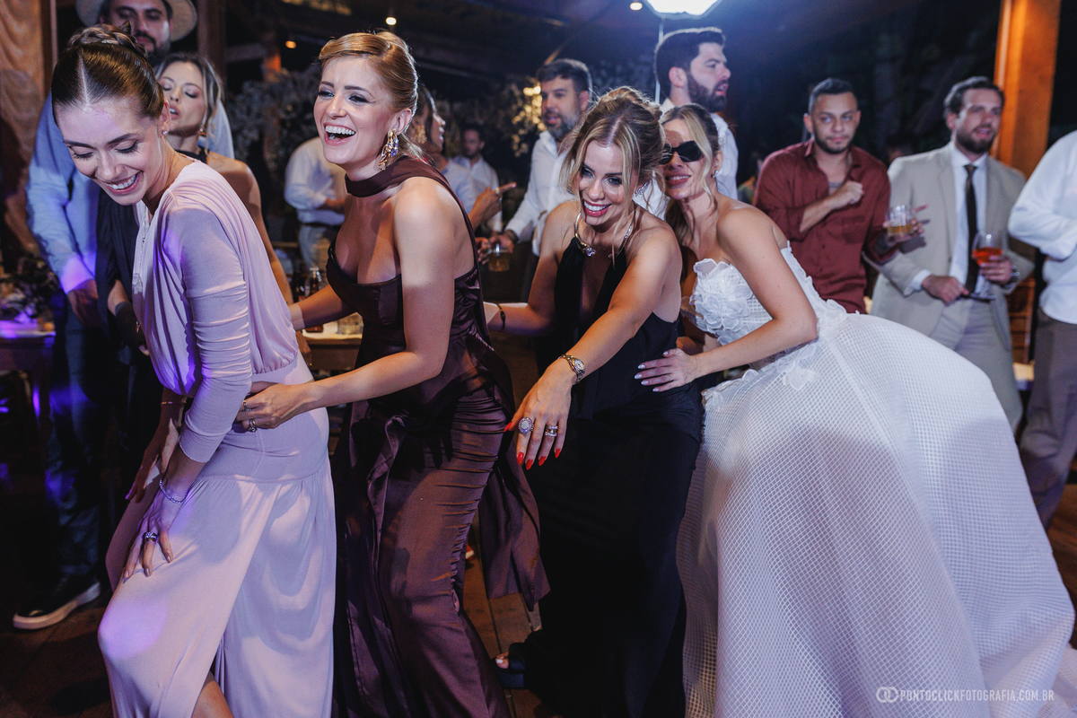 Noiva e convidadas dançam juntas em fila, sorrindo na pista da festa de casamento. Fotógrafo de casamento Pontoclick Fotografia.