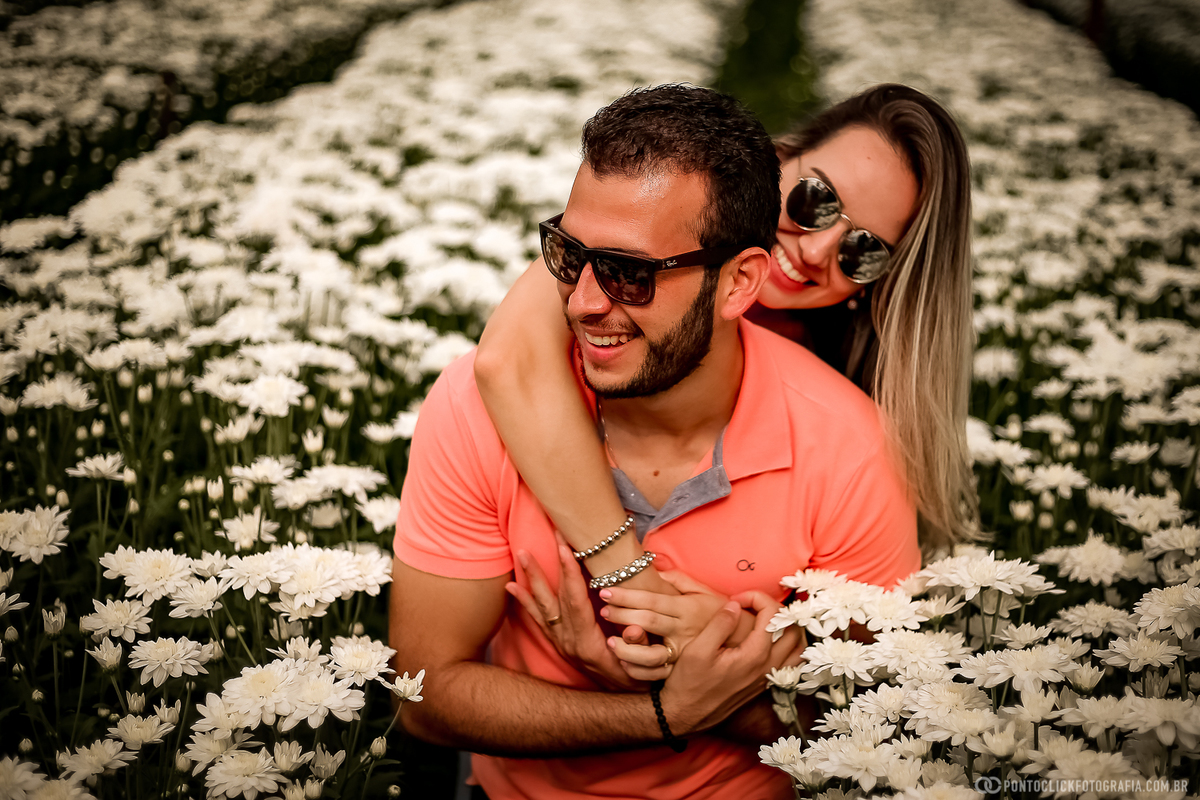 noiva abraçando o noivo com a mão por cima do ombro dele eles estão de oculos de sol e sorrindo muito no meio da plantação de flores em holambra