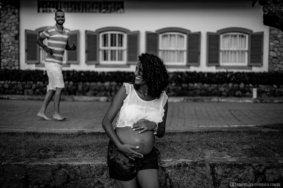gestante sentada com as duas maos na barriga e sorrindo e o papai corrnendo e sorrindo ao fundo