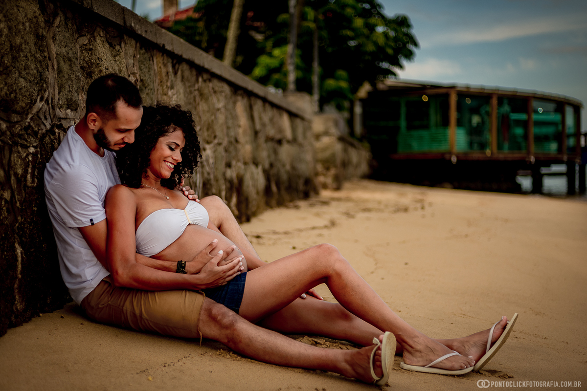 casal sentado na area da praia em ilhabela segurando a barriga da mamae