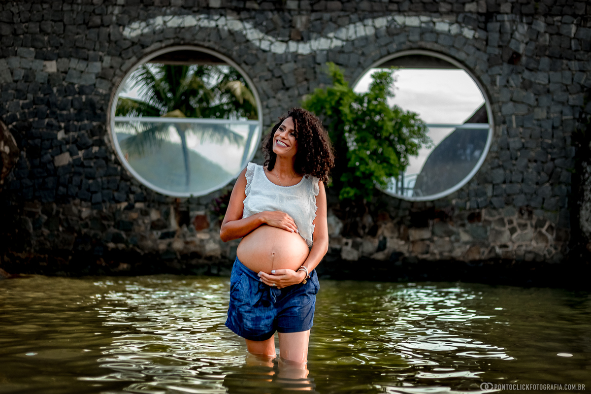 mamae gestante segurando a barriga e sorrindo dentro da agua na praia em ilhabela