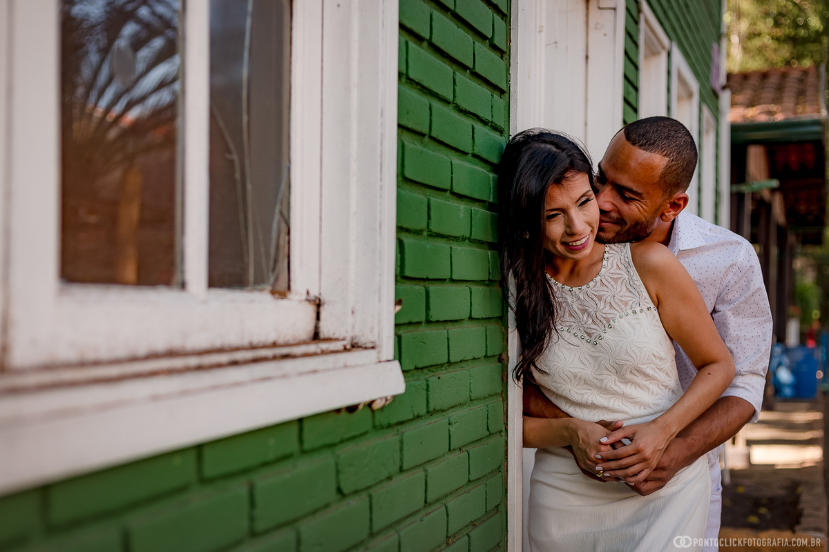 noivo dando um cheiro no pescoço da sua noiva e abraçados e encostados num muro verde em Holambra estão vestidos de branco