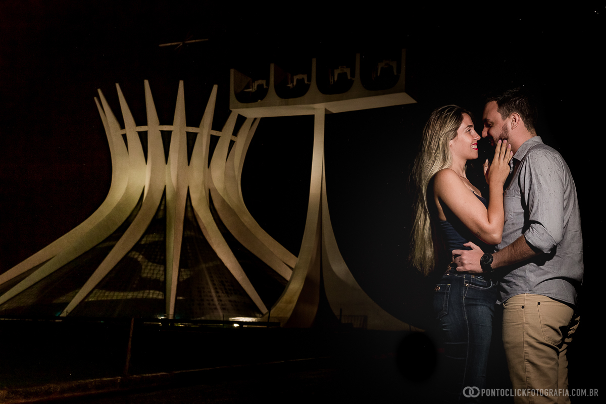 casal abraçado e noiva com a mao no rosto do noivo e ao fundo a catedral de brasilia em ensaio fotografico