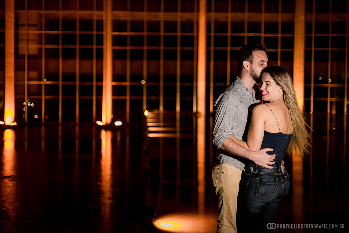 casal abraçado e noiva de costa sorrindo com o noivo beijando no rosto dela em ensaio fotografico noturno em brasilia