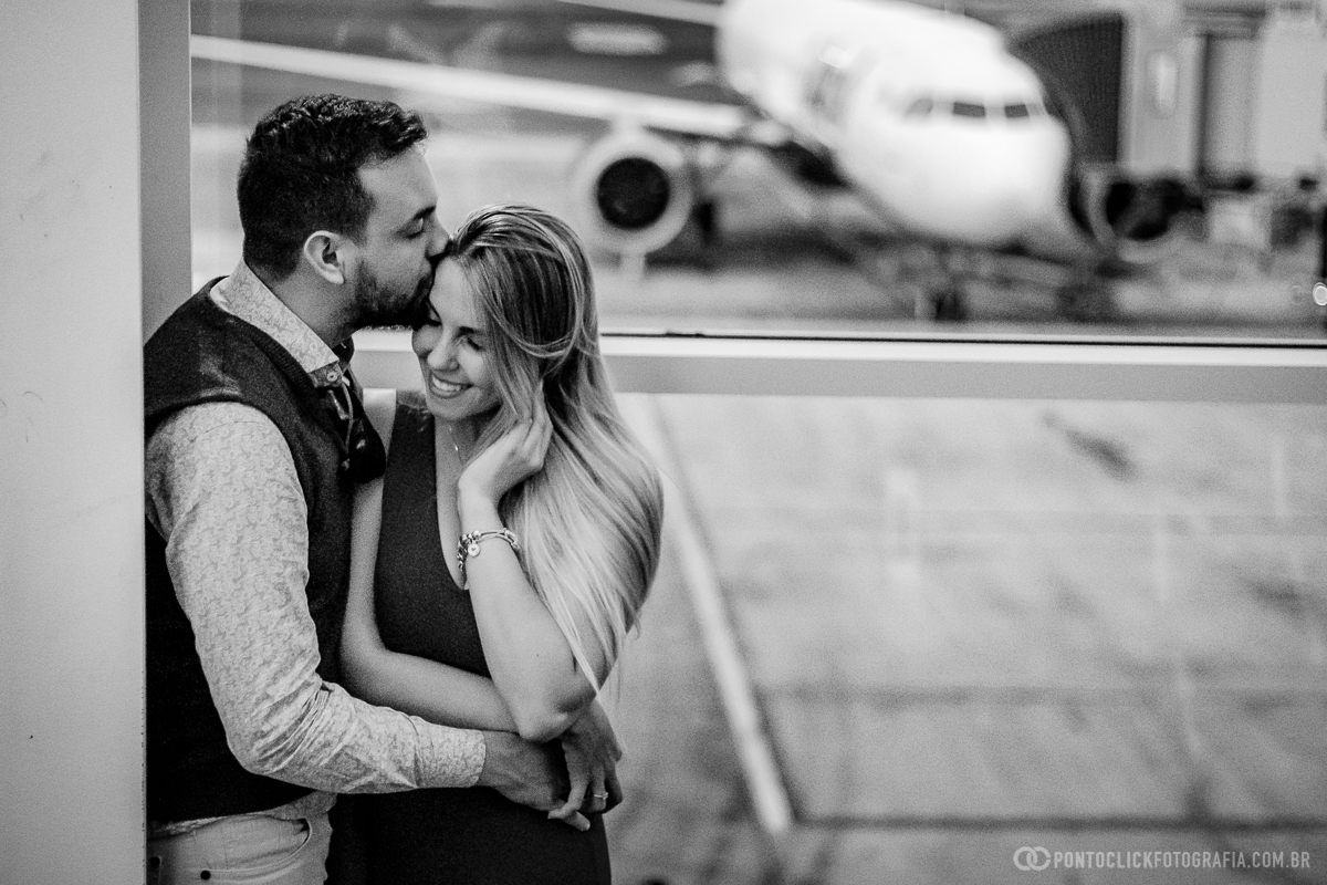 casal abraçados e a noiva sorrindo encostados na parede e no fundo um aviao estacionado em ensaio fotografico em ensaio fotografico no aeroporto
