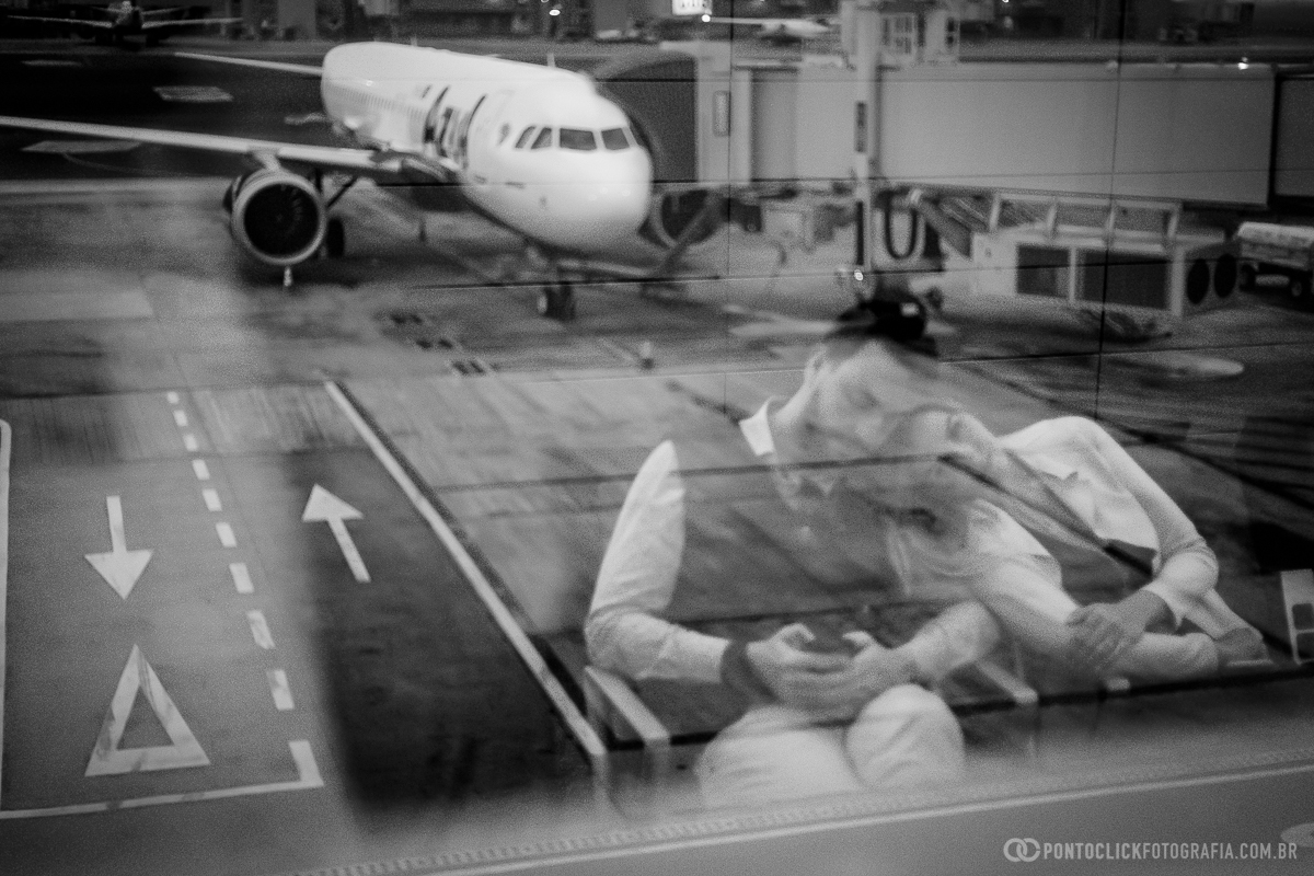 aviao no fundo estacionado do aeroporto de viracopos  e o reflexo do casal no vidro com a noiva deita no ombro do noivo em ensaio fotografico no aeroporto