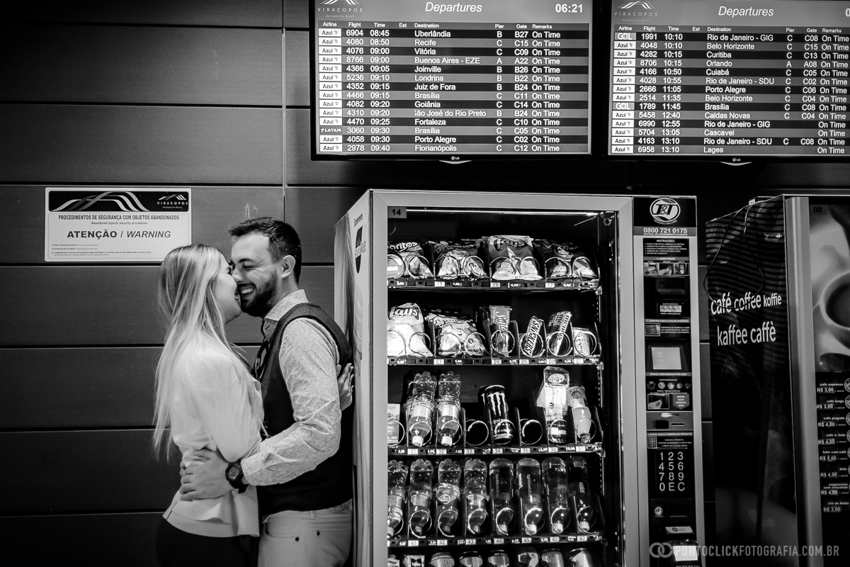 casal abraçado e sorrindo encostados em uma maquina de lanches no salao de embarque do aeroporto de viracopos em ensaio fotografico no aeroporto