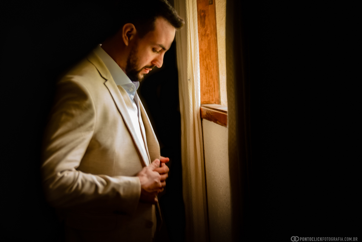 noivo abotoando o terno de casamento na frente da janela do quarto do noivo do espaço laforet em sao roque são paulo