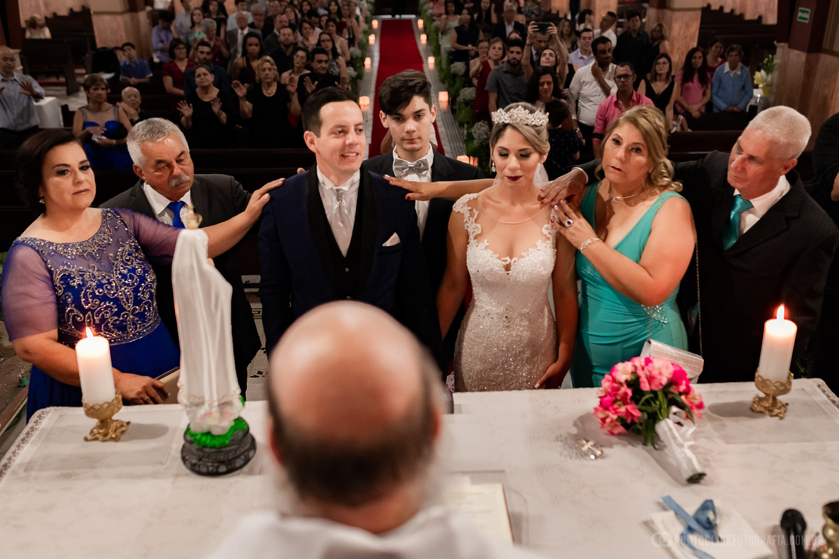 padre da igreja da matriz de sao roque mae com vestido azul escuro com a mao no ombro do noivo pae do noivo com terno preto com a mao no ombro no noivo noivo com terno azul filho do casal ao fundo noiva com vestido brando recebendo a bencao da sua mae 