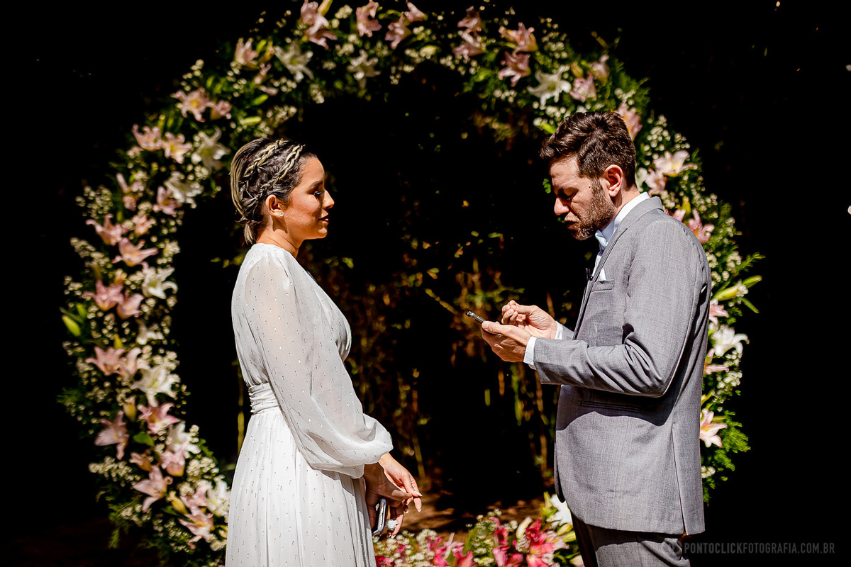 noivo lendo os votos para a noiva e noiva olhando para ele e ao fundo um lindo arco de flores no espaco lafort sao roque fotos de casamento