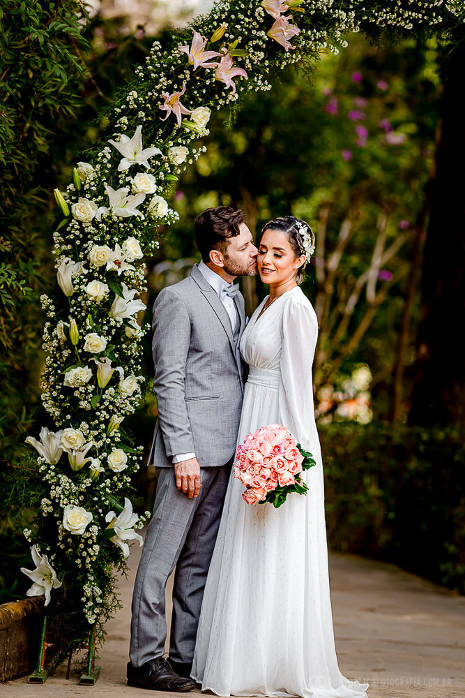 noivo beijando a noivo no rosto e noiva segurando o buquet e debaixo do arco de flores no Elopement Wedding no Espaço Laforet