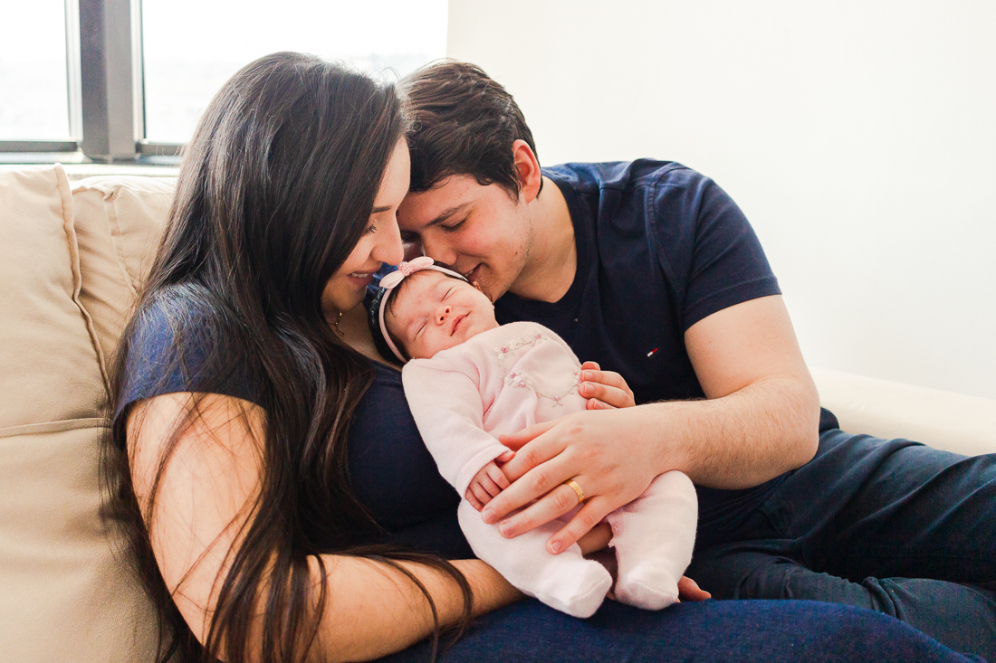 Foto dos pais com filha no colo dando beijo e carinho no ensaio em casa realizado na zona leste da cidade de São Paulo