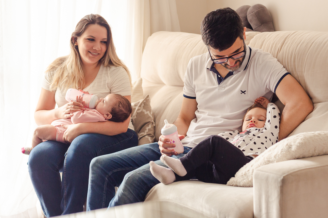 Foto dos pais com filhas no colo dando mamadeira no ensaio lifestyle de família realizado na cidade de São Paulo