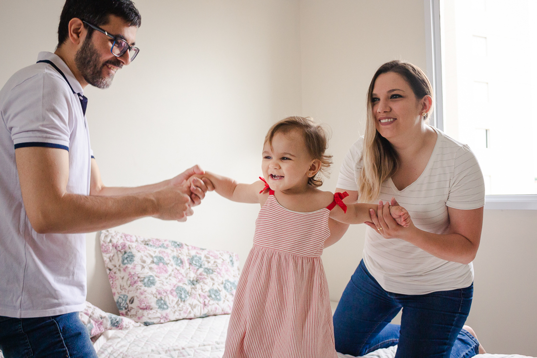 Foto de pais de menina brincando no quarto dos pais em casa, no ensaio de família lifestyle em São Paulo