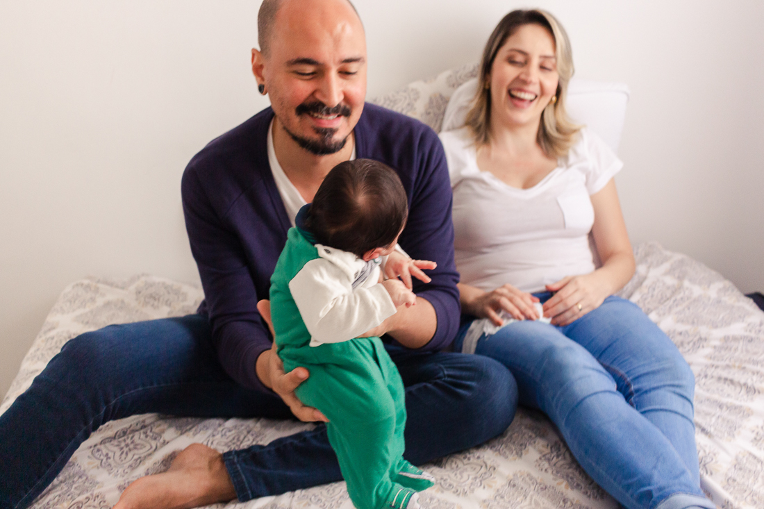 Pais sentados na cama e sorrindo segurando o bebe no colo dentro do quarto no ensaio lifestyle de família na cidade de SP