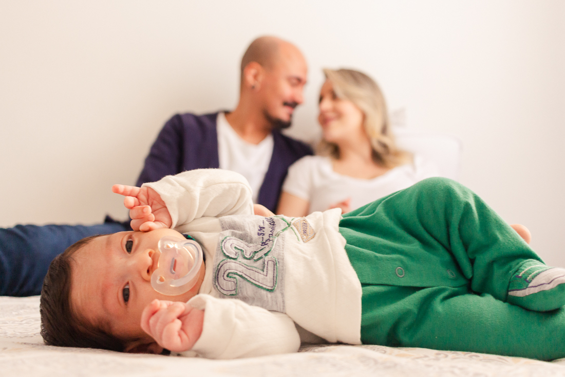 Pais sentados na cama e sorrindo com bebe deitado na cama dentro do quarto no ensaio lifestyle de família na cidade de SP