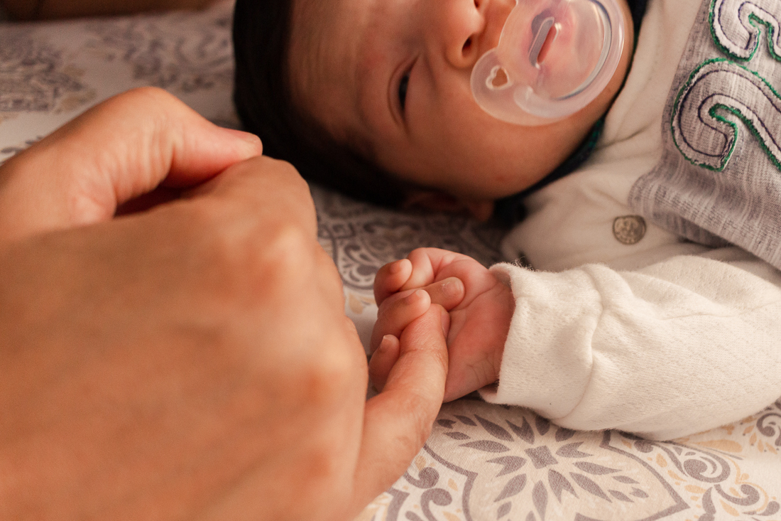 Detalhes do bebe dormindo na cama com  as mãos juntas, com o bebe segurando a o dedo do pai de primeira viagem.