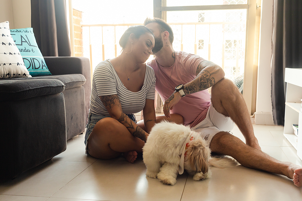 Foto do casal no chão da sala de casa se beijando e fazendo carinho no cachorrinho, no ensaio lifestyle na cidade de SP