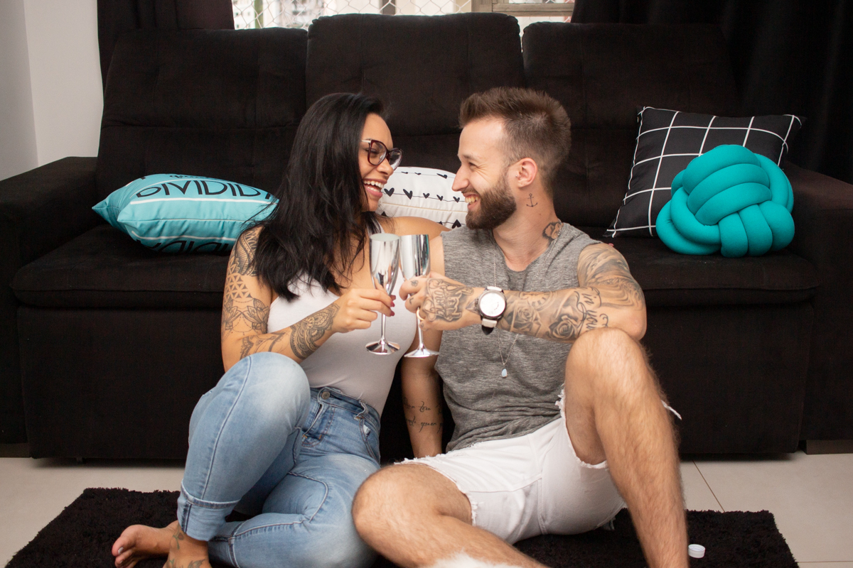Foto do casal no chão da sala de casa, comemorando com taça de champanhe e vinho no ensaio lifestyle na cidade de SP