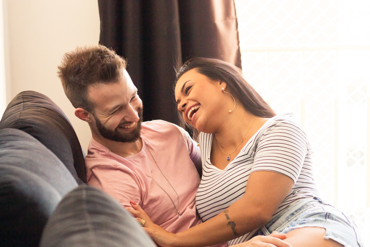 Foto do casal no sofá de casa sorrindo espontaneamente e fazendo carinho, no ensaio lifestyle no bairro Vila Ema na cidade de SP